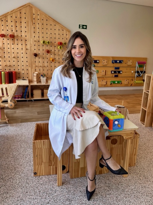 Dra. Yasmin Miglio sorrindo, sentada na sala de atendimento infantil do consultório.